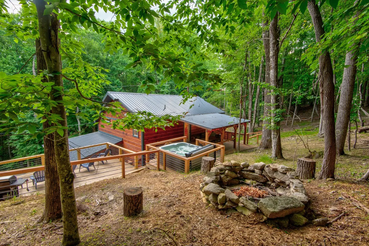 This exterior shot showcases the log cabin structure, an inviting hot tub set on the deck, a cozy fire pit area, and comfortable deck seating. The surrounding natural landscape creates a tranquil and rejuvenating atmosphere, with ample space for group gatherings and relaxation.