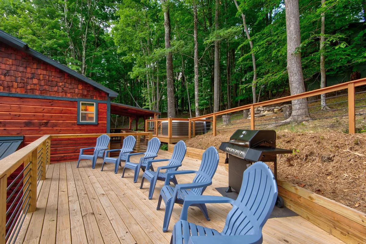 This image focuses on the spacious wooden deck with bright blue Adirondack chairs, a gas grill ready for barbecues, and lush trees surrounding the area. The viewpoint invites the viewer to picture themselves enjoying a meal or relaxing while taking in the sunset or stars.
