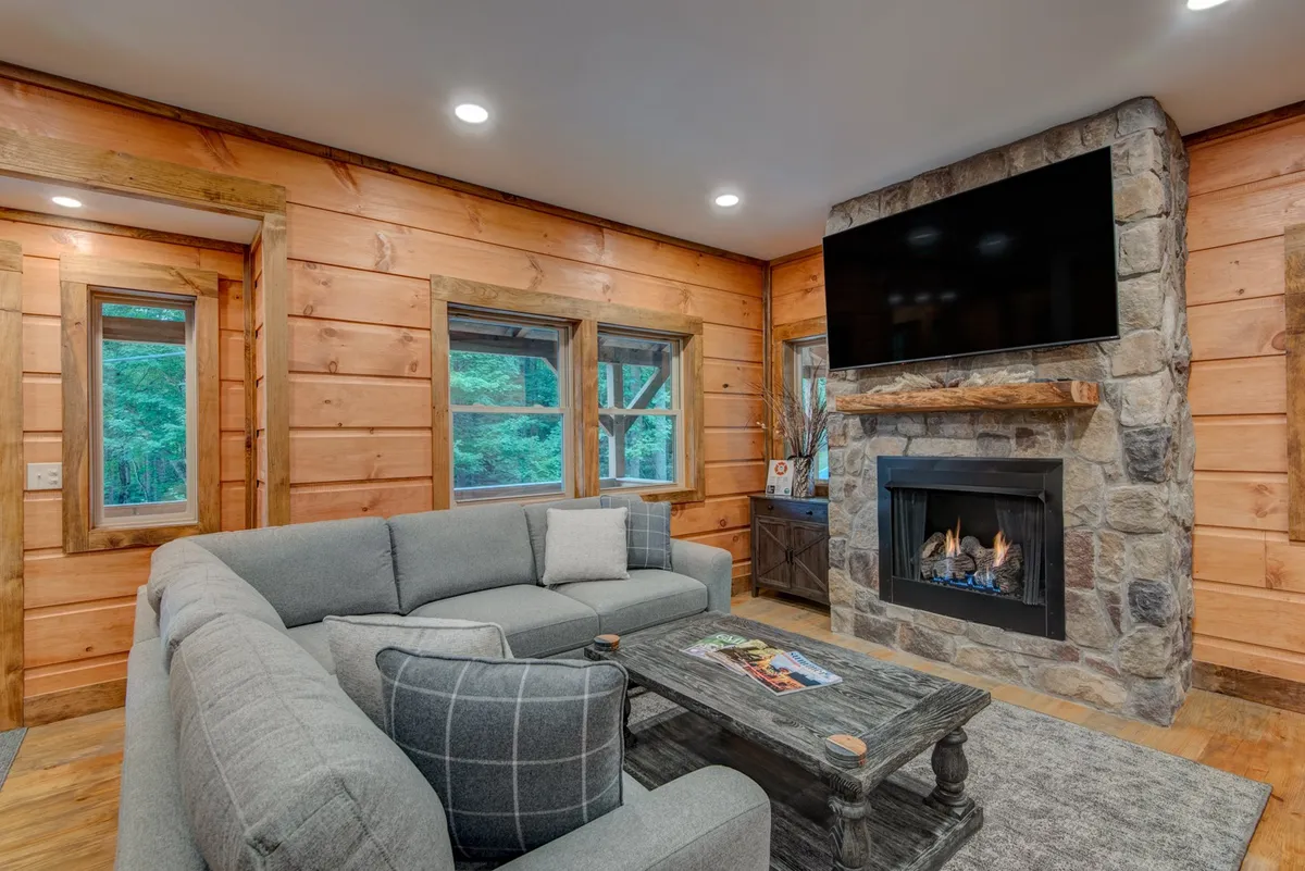 A close-up of the living room's centerpiece: a plush sectional sofa positioned in front of a prominent stone fireplace, with a large flatscreen TV mounted nearby and a rustic wooden coffee table completing the inviting setting. The arrangement suggests a perfect spot for cozy movie nights or warming up after outdoor adventures.