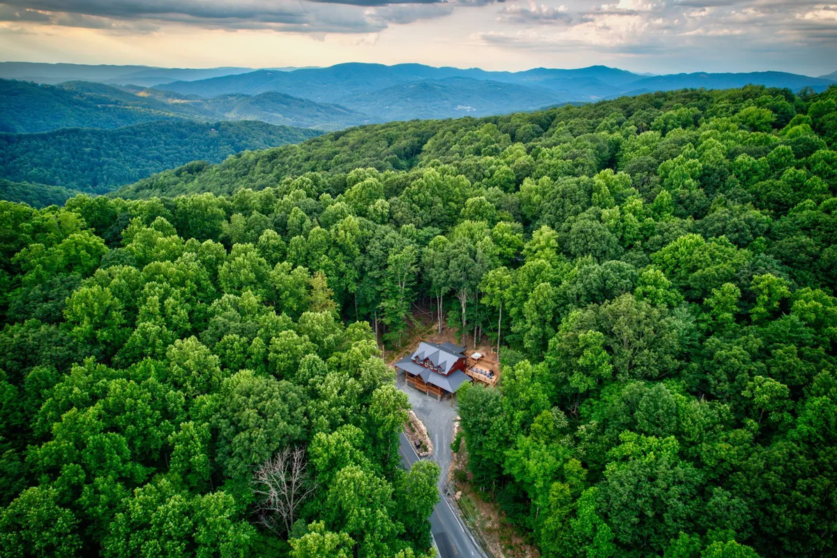 A classic wooden cabin nestled among lush trees, with a winding road leading toward the mountains in the distance. The setting evokes a sense of adventure and seclusion, with the landscape hinting at outdoor activities like hiking, skiing, and exploring nearby trails.