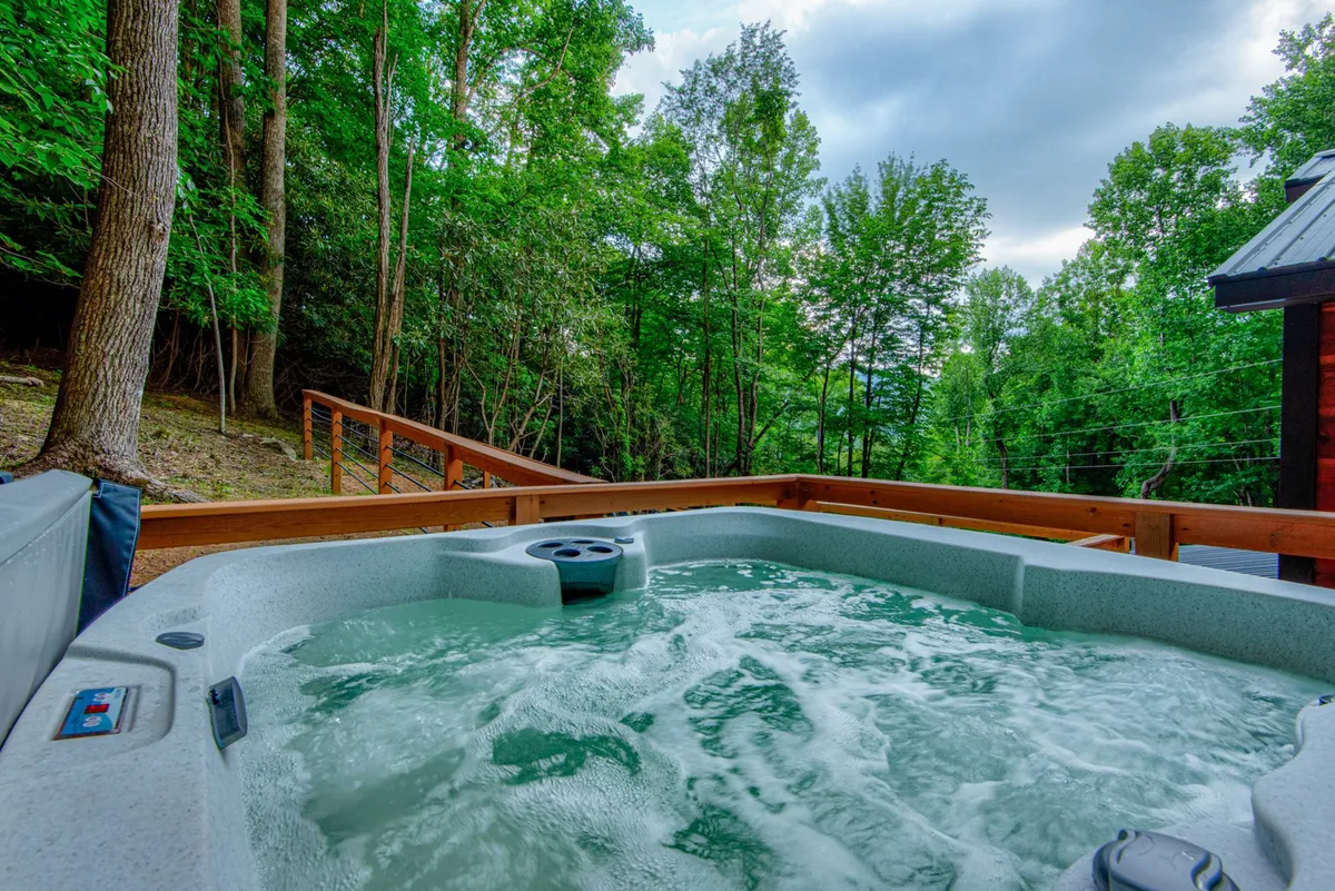 A tranquil close-up of the bubbling hot tub nestled on the wooden deck, framed by green trees. The intimate setting is ideal for relaxing soaks after a day of adventure, with the promise of stargazing above.