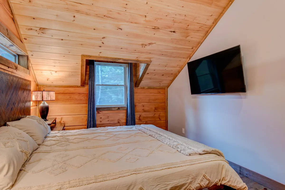 A bedroom with a comfortable bed, a window allowing in daylight, a bedside lamp, and a flatscreen TV. The mood is comforting, ideal for relaxing, reading, or catching up on favorite shows before bed.