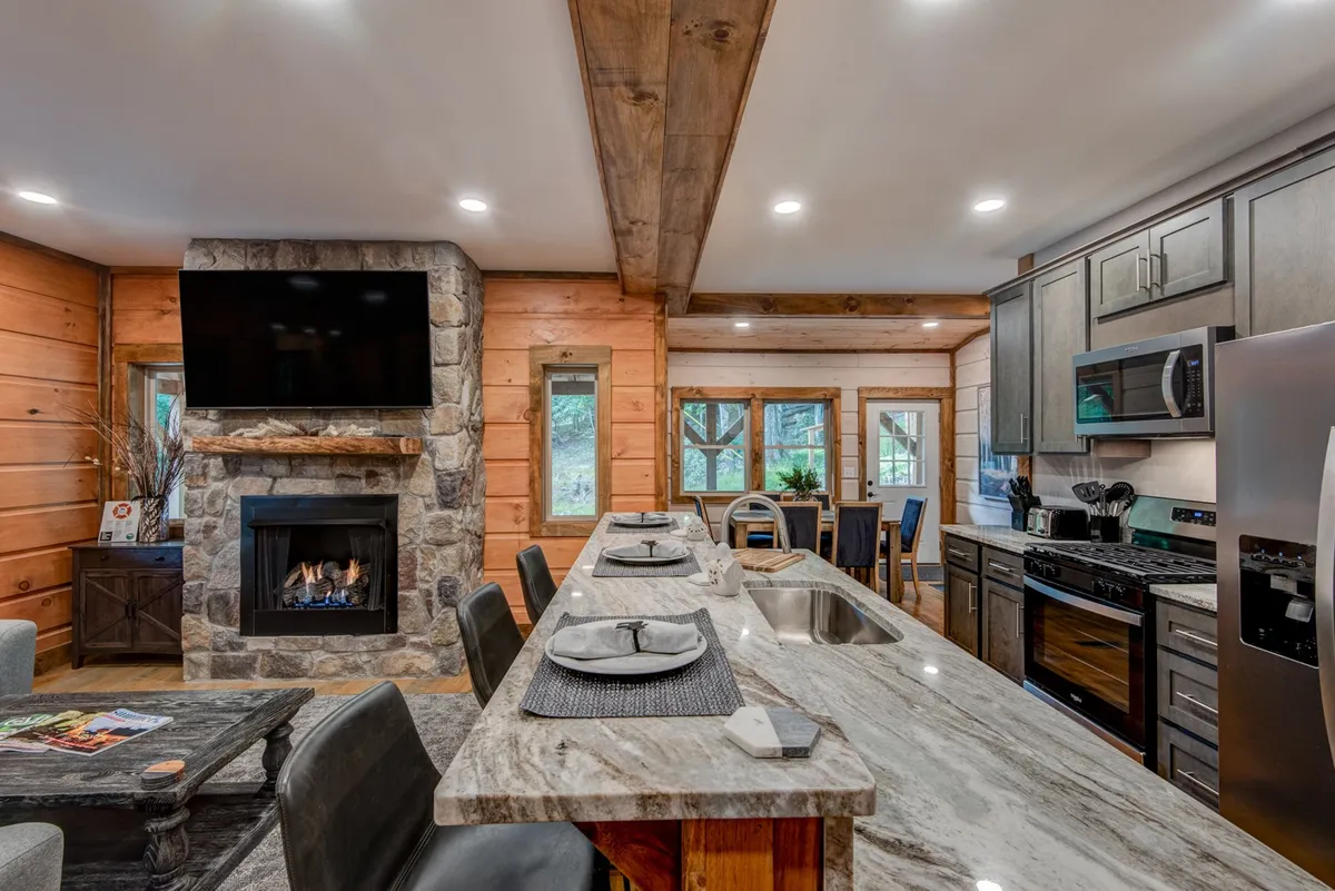 A wide-angle view of the main interior gathering space, capturing the open-concept living area with a plush sectional facing a stunning stone gas log fireplace, granite countertops stretching into a modern kitchen with high-end appliances, and a dining area adjacent to large windows. The warm lighting and thoughtful layout convey a sense of comfort and togetherness, inviting both relaxation and socializing.