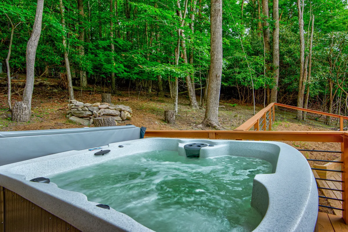 This exterior image focuses on a private hot tub nestled on a wooden deck, surrounded by lush trees and enclosed by a deck railing. The tranquil setting hints at relaxation and the opportunity to soak while enjoying scenic views, with nature as the backdrop.