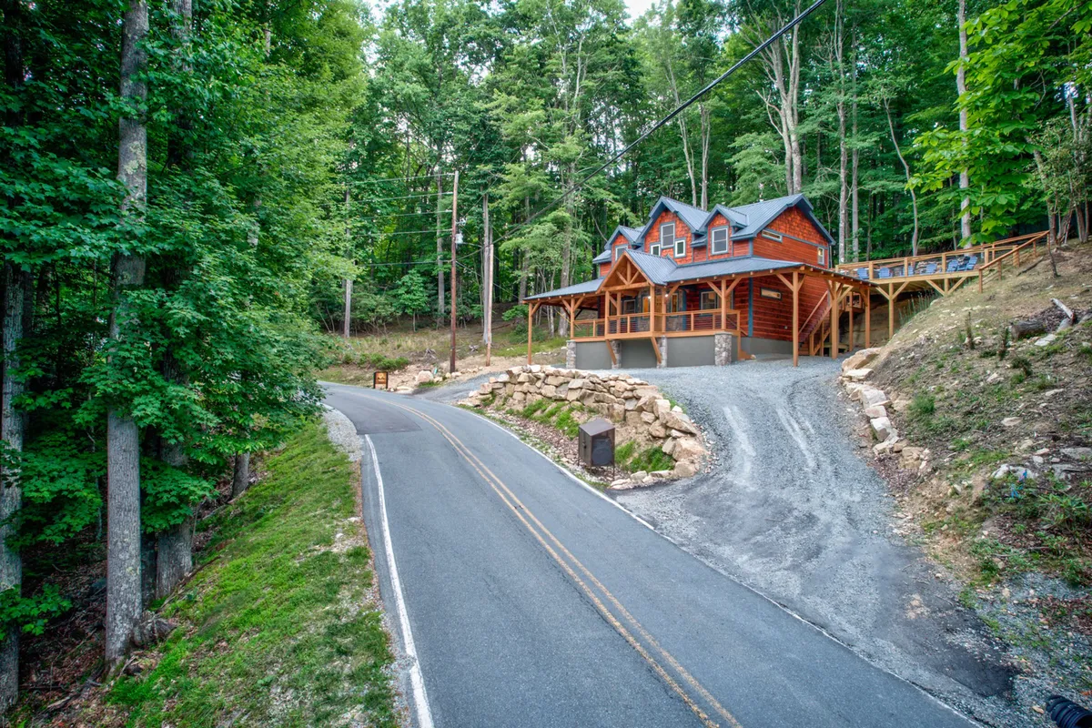 A welcoming view of the cabin set among tall trees, with a winding road leading up to the home. The porch invites guests to gather, and the surrounding natural landscape hints at outdoor adventures and evenings spent under the stars.