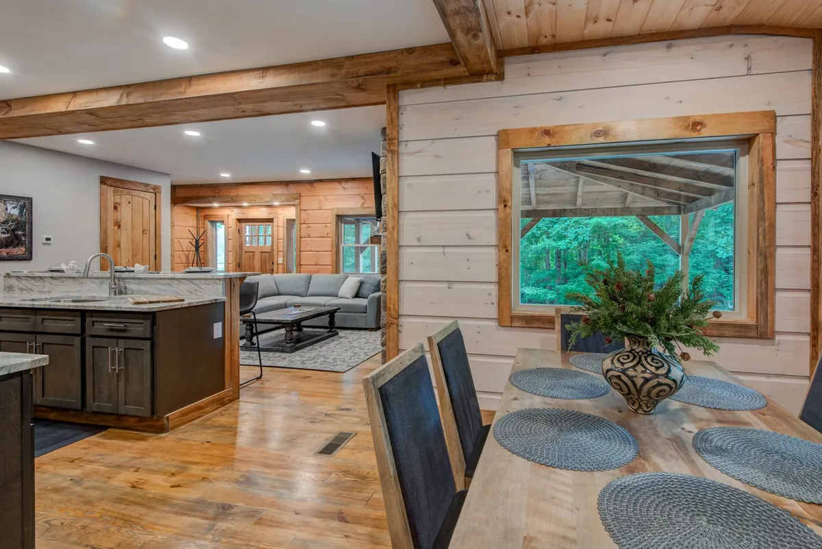 A perspective from the kitchen island looking into the living space, with stylish bar seating, a glimpse of the granite countertop, and the living area framed by a large window that provides mountain views. The setup suggests morning coffee, group meals, or casual conversations, with natural light enhancing the warm, communal atmosphere.